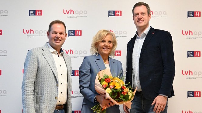 Home Drei Personen posieren vor einem Hintergrund mit Logos, eine hält einen Blumenstrauß